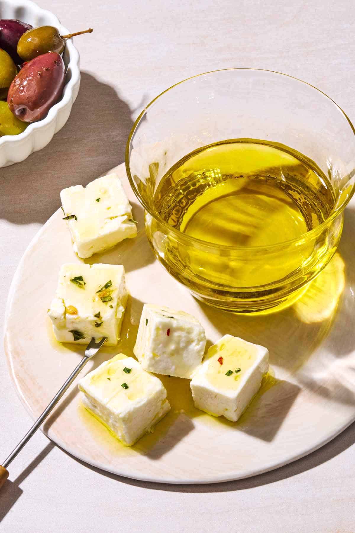 A close up of a clear bowl of olive oil on a plate with cubes of feta cheese. Behind this is a bowl of olives.