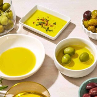 A close up of a bowl of green olives, a bowl of kalamata olives, a bowl of mixed olives, and 3 bowls of olive oil, one with 3 green olives in it.