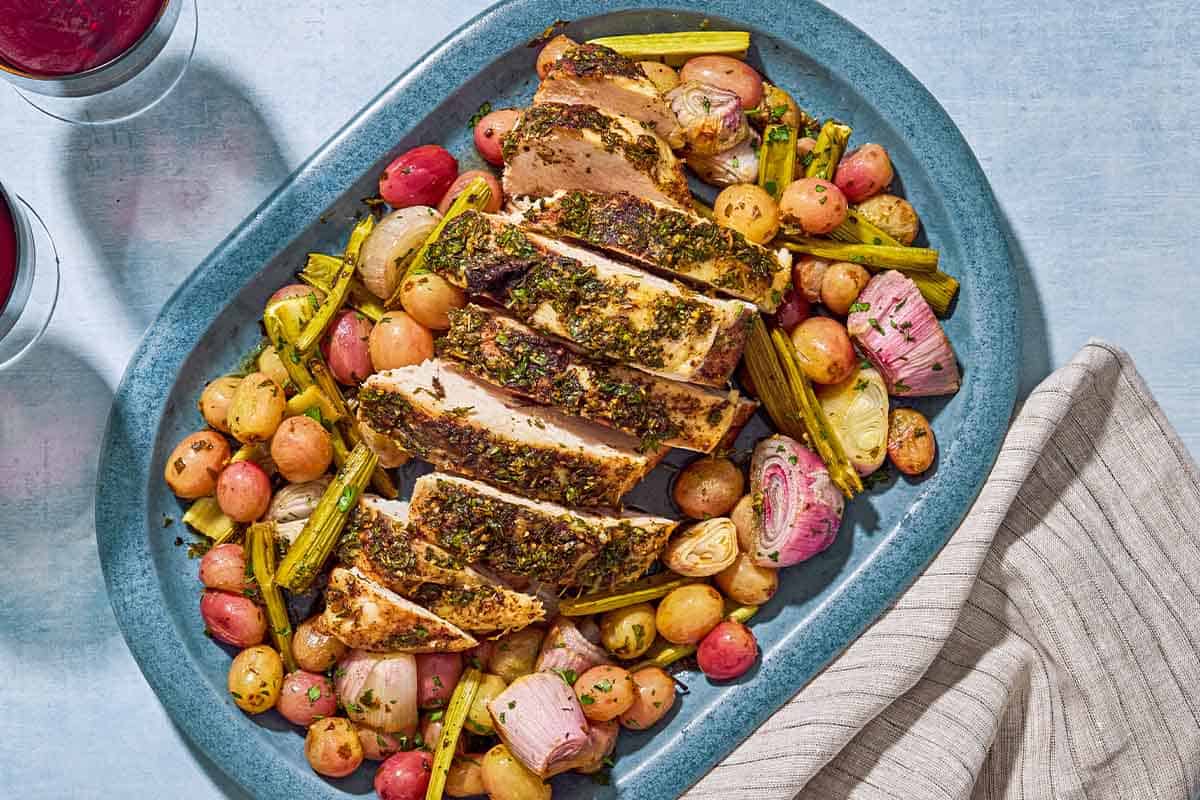 An overhead photo of a sliced roast turkey breast with grapes and vegetables on a serving platter. Next to this is a cloth napkin and glasses of red wine.