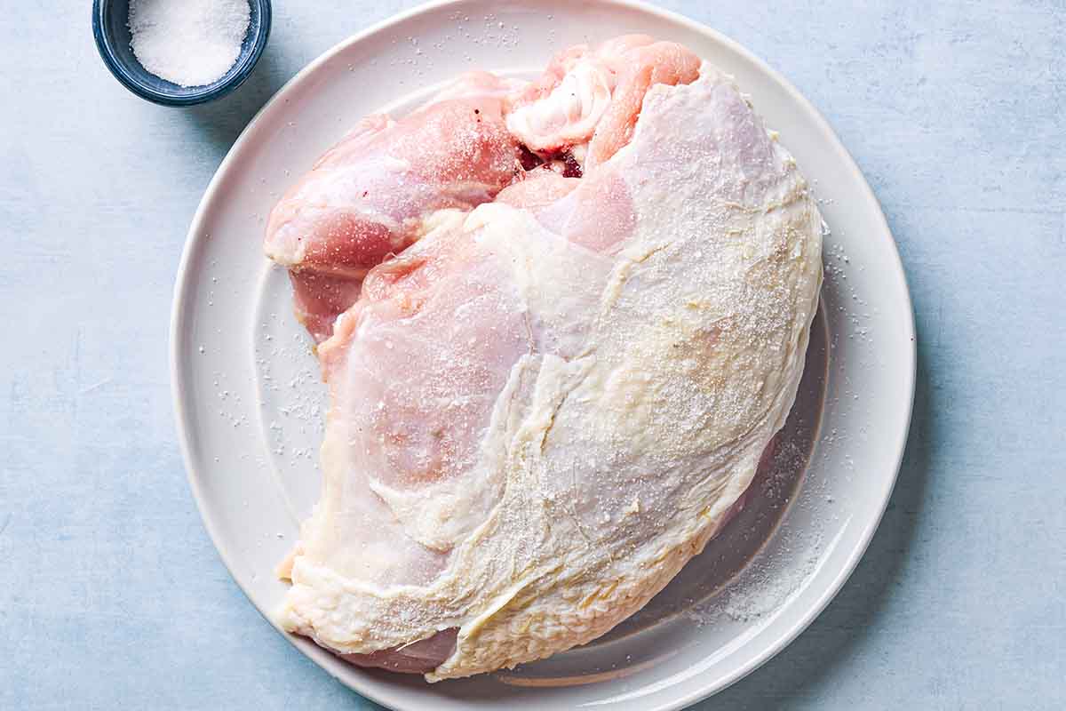 An overhead photo of an uncooked, bone-in turkey breast seasoned with salt on a plate. Next to this is a small bowl of salt.