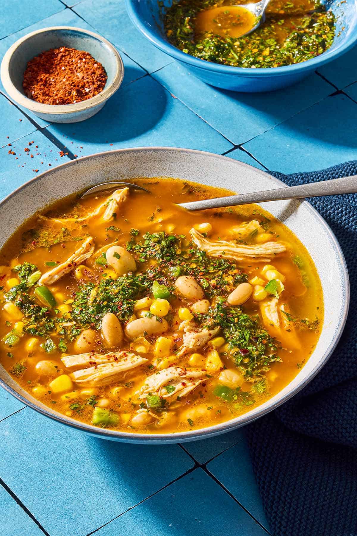 White chicken chili topped with chermoula in a bowl with a spoon. Next to this is a kitchen towel, a bowl of aleppo pepper and a bowl of chermoula with a spoon.