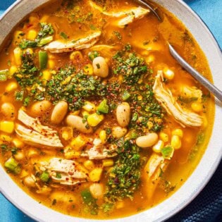 An overhead photo of white chicken chili topped with chermoula in a bowl with a spoon. Next to this is a kitchen towel.