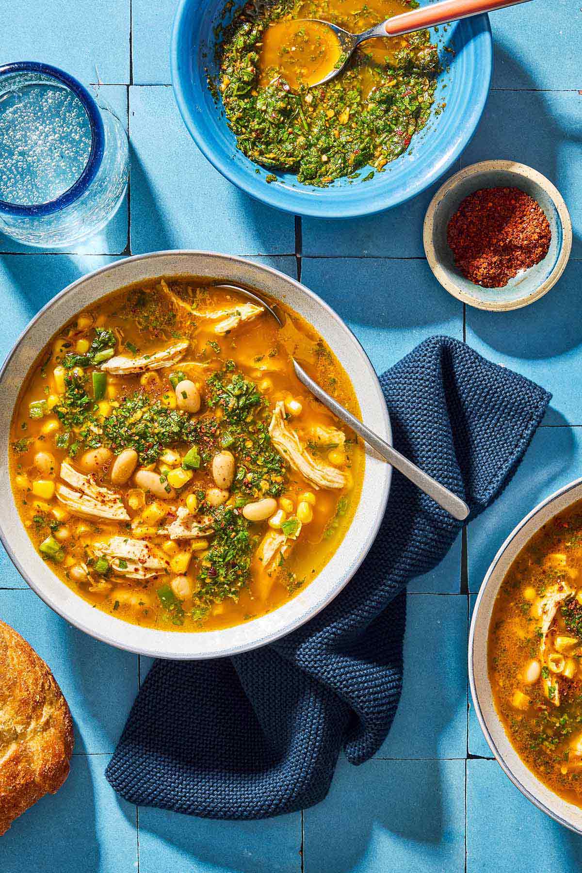 An overhead photo of white chicken chili topped with chermoula in a bowl with a spoon. Next to this is a kitchen towel, another bowl of the soup, a glass of water, a bowl of aleppo pepper and a bowl of chermoula with a spoon.