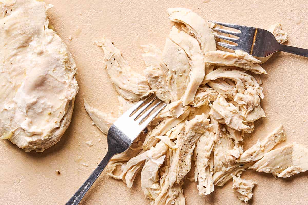 An overhead photo of cooked chicken being shredded with 2 forks.