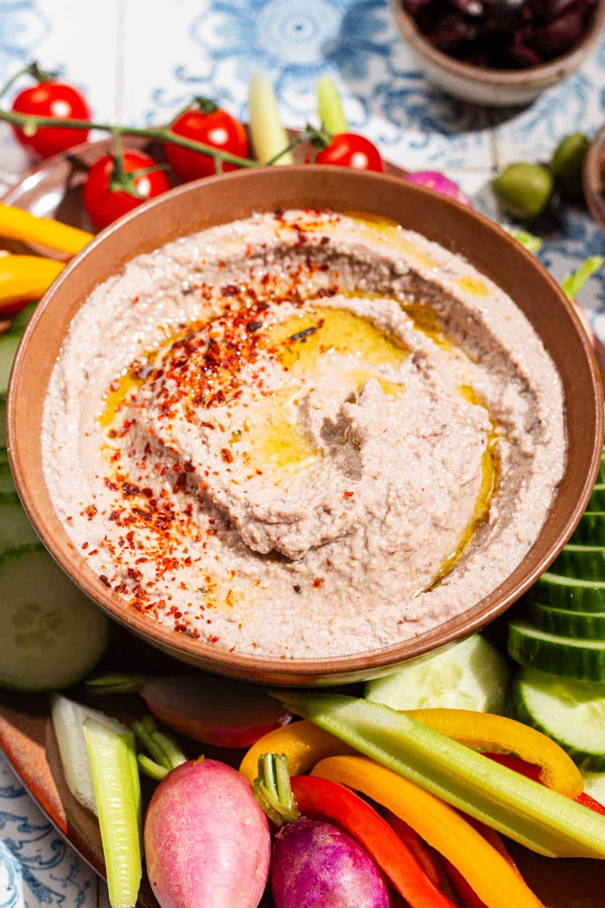 A close up of a tarator sauce in a bowl garnished with olive oil and aleppo pepper. Surrounding this is a variety of raw vegetables.