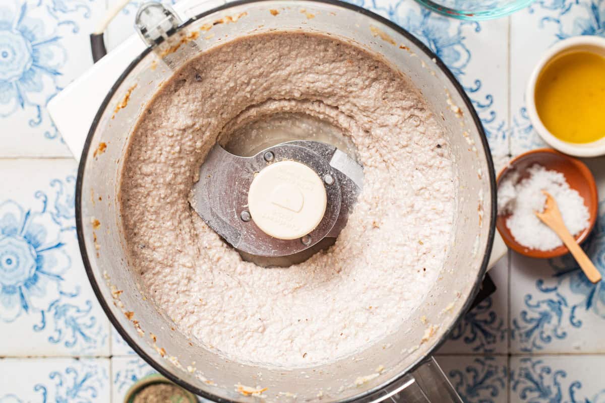 An overhead photo a paste made with bread, garlic, walnuts, and lemon juice in the bowl of a food processor fitted with a blade. Next to this are small bowls of olive oil and salt.