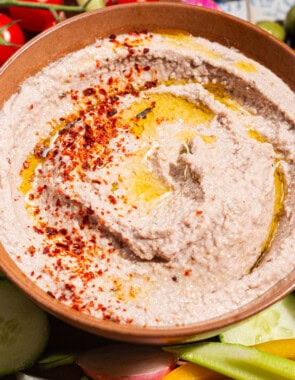 A close up of a tarator sauce in a bowl garnished with olive oil and aleppo pepper. Surrounding this is a variety of raw vegetables on a platter.