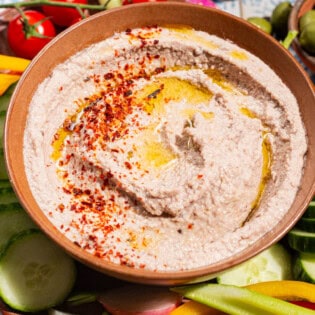 A close up of a tarator sauce in a bowl garnished with olive oil and aleppo pepper. Surrounding this is a variety of raw vegetables on a platter.