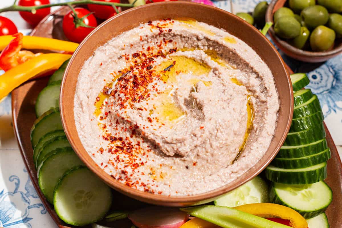 A close up of a tarator sauce in a bowl garnished with olive oil and aleppo pepper. Surrounding this is a variety of raw vegetables on a platter.