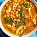 An overhead photo of white chicken chili topped with chermoula in a bowl with a spoon. Next to this is a kitchen towel.