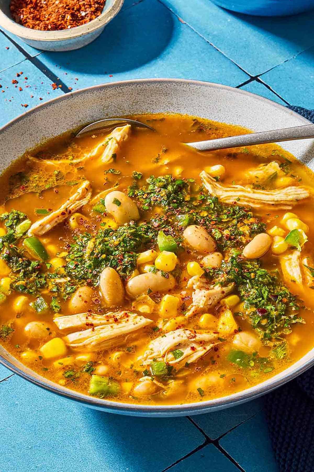 A close up of white chicken chili topped with chermoula in a bowl with a spoon. Next to this is a bowl of aleppo pepper.