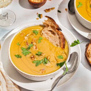 A close up of acorn squash soup garnished with walnuts, parsley and a drizzle of olive oil in a bowl with a piece of bread. Next to this is a spoon, a cloth napkin, another bowl of the soup, a glass of white wine, some parsley and small bowls of walnuts and salt.