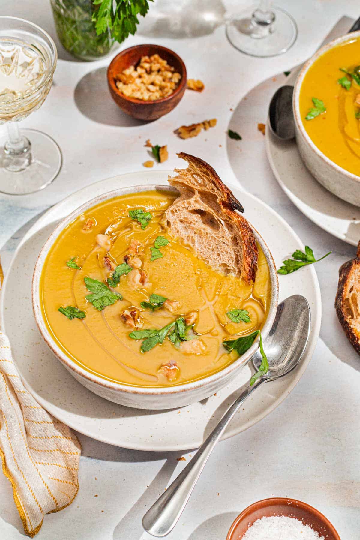 A close up of acorn squash soup garnished with walnuts, parsley and a drizzle of olive oil in a bowl with a piece of bread. Next to this is a spoon, a cloth napkin, another bowl of the soup, a glass of white wine, some parsley and small bowls of walnuts and salt.