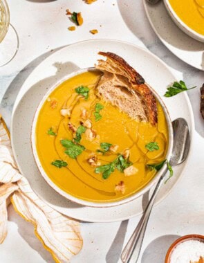 An overhead photo of acorn squash soup garnished with walnuts, parsley and a drizzle of olive oil in a bowl with a piece of bread. Next to this is a spoon, a cloth napkin, another bowl of the soup, and small bowls of walnuts and salt.