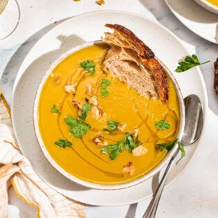 An overhead photo of acorn squash soup garnished with walnuts, parsley and a drizzle of olive oil in a bowl with a piece of bread. Next to this is a spoon, a cloth napkin, another bowl of the soup, and small bowls of walnuts and salt.