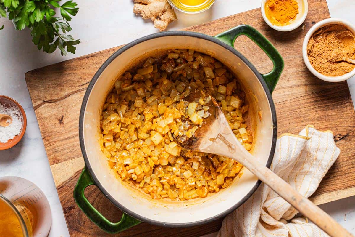 An overhead photo of chopped onion with garlic, ras el hanout, turmeric and salt in a large pot with a wooden spoon after being sauteed. Next to this is a cloth napkin, ginger root, a bit of parsley, vegetable broth, and small bowls of salt, olive oil, turmeric and ras el hanout.