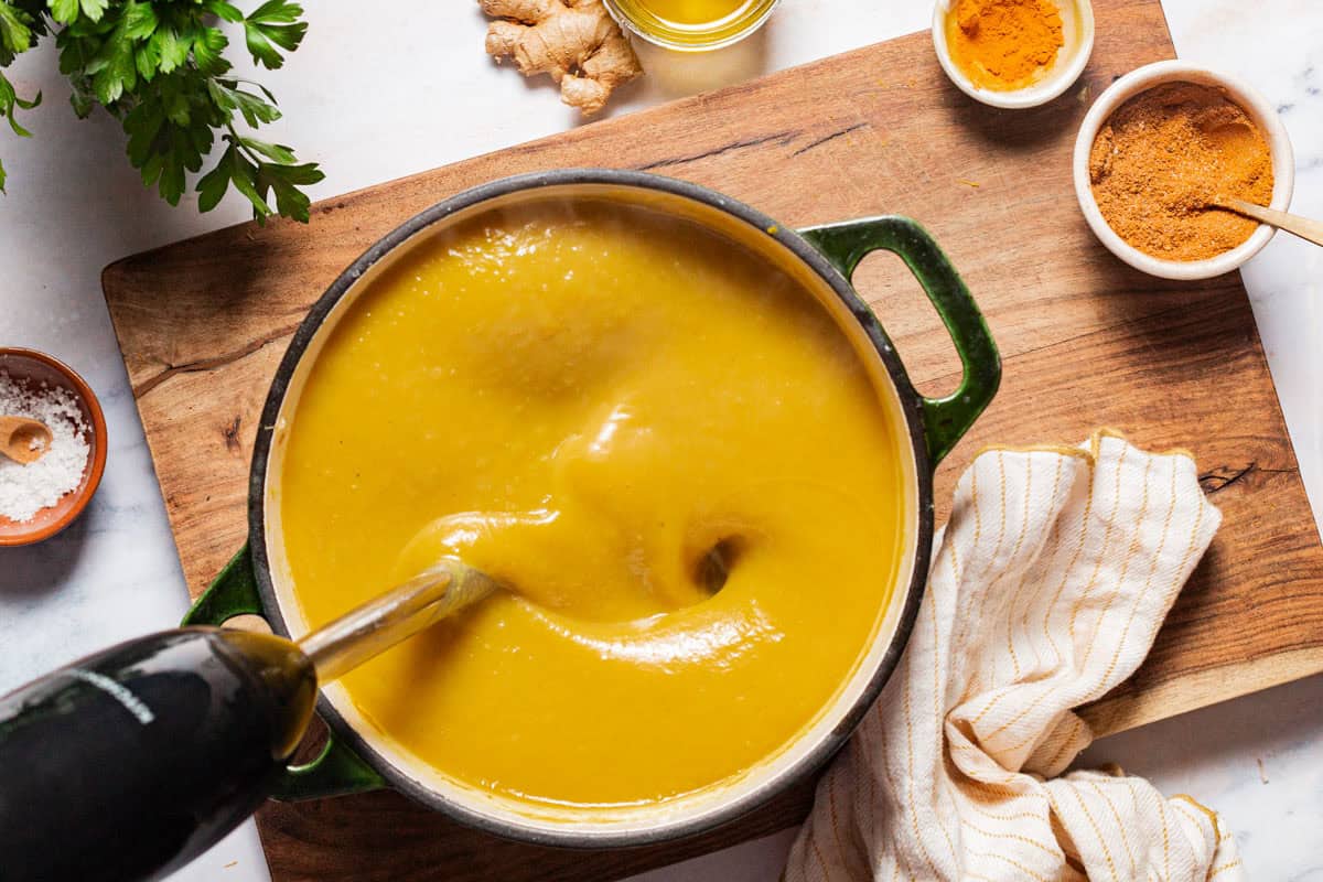 An overhead photo of acorn squash soup in a large pot being blended with an immersion blender. Next to this is a cloth napkin, a piece of ginger root, a bit of parsley, and small bowls of salt, olive oil, turmeric and ras el hanout.