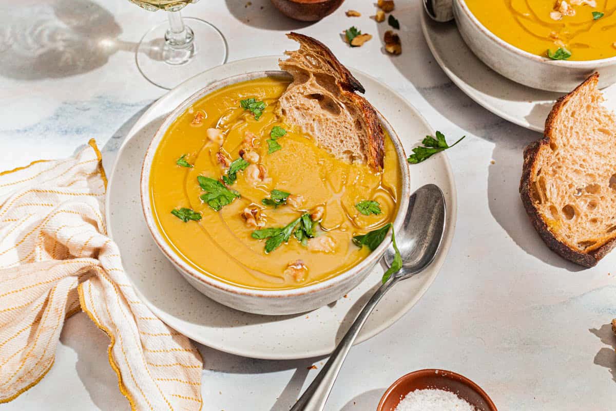 A close up of acorn squash soup garnished with walnuts, parsley and a drizzle of olive oil in a bowl with a piece of bread. Next to this is a spoon, a cloth napkin, another bowl of the soup with a slice of bread, and a small bowl of salt.