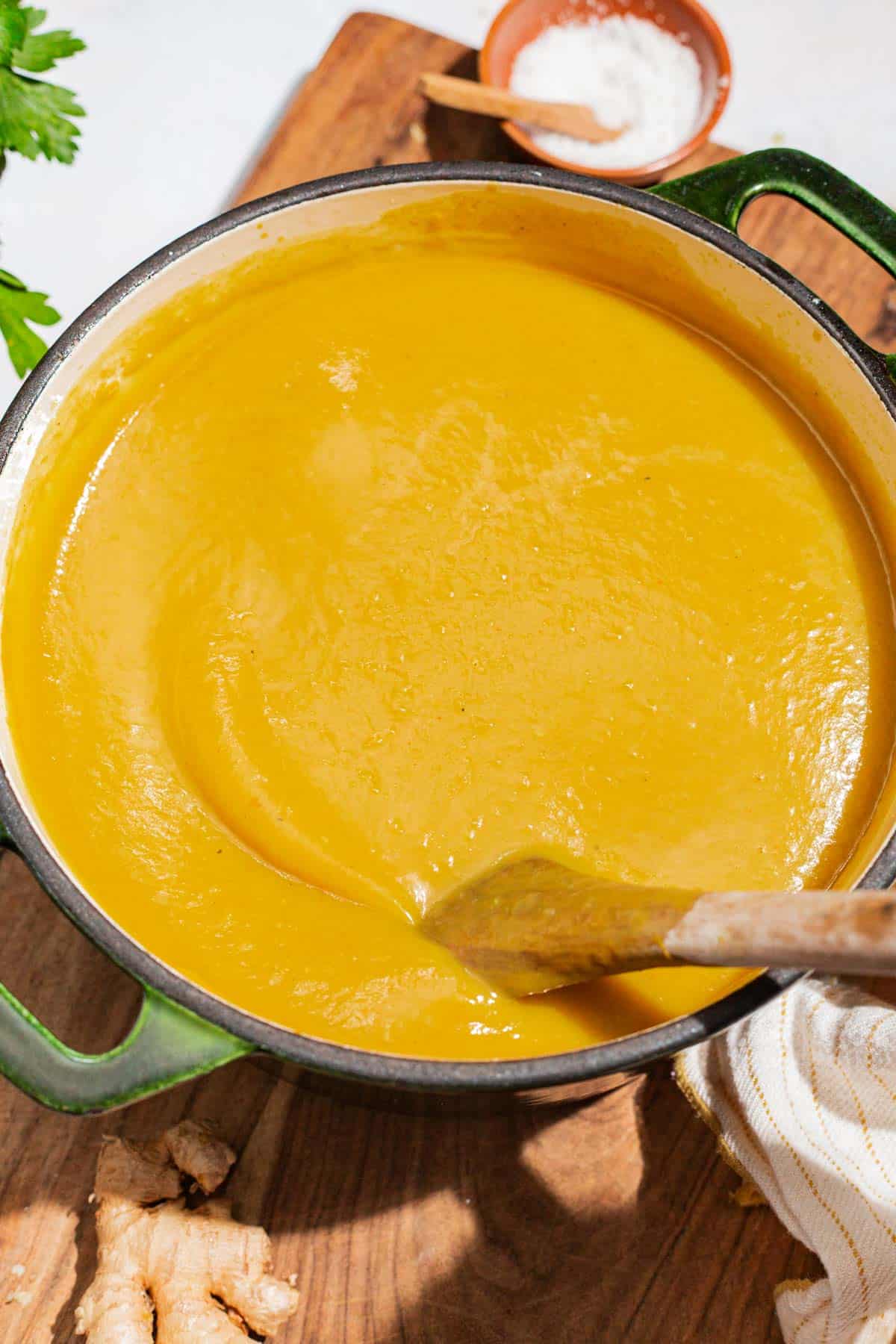 A close up of acorn squash soup in a large pot with a wooden spoon. Next to this is a cloth napkin, a small bowl of salt, and a piece of ginger root.