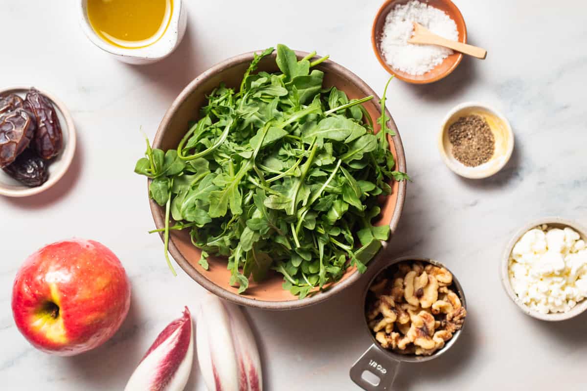 Ingredients for apple walnut salad including endive, baby arugula, pitted dates, an apple, goat cheese, apple cider vinaigrette, salt, and black pepper.