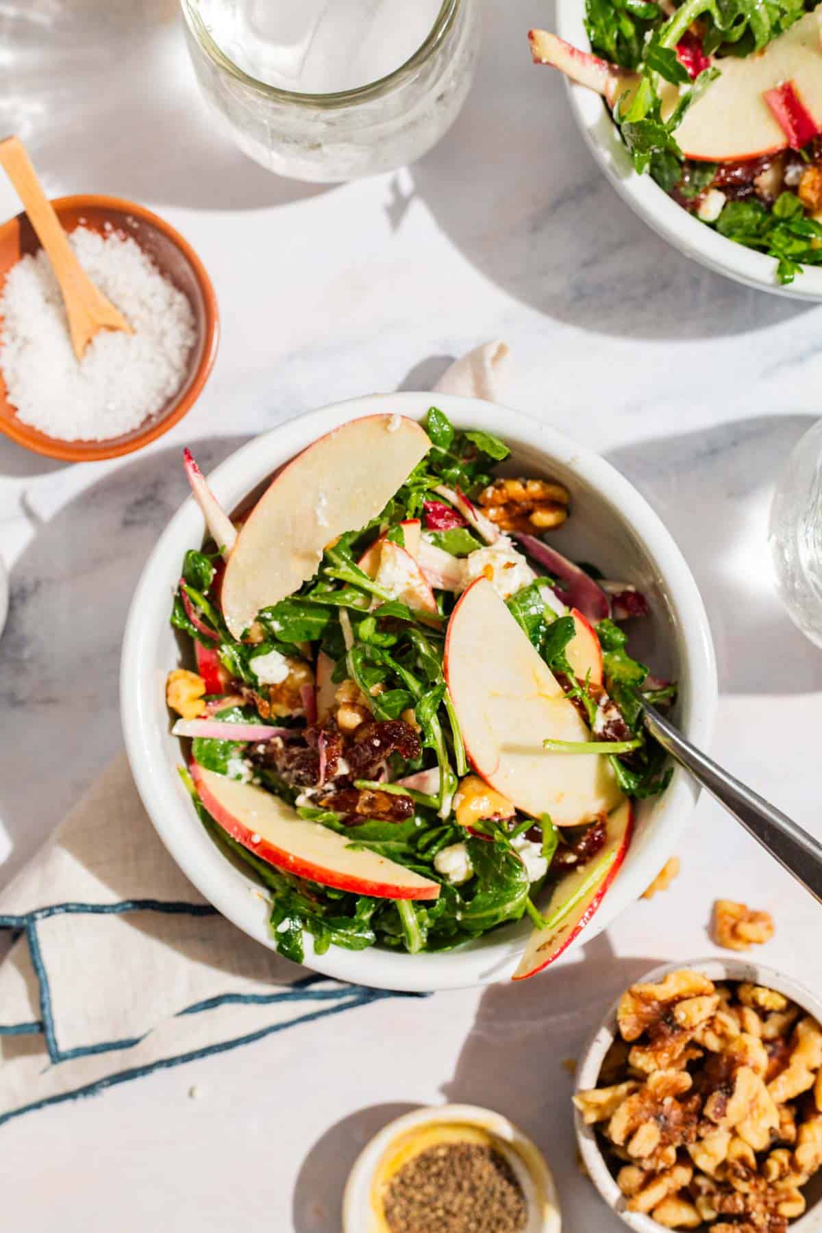 An overhead photo of a serving of the apple walnut salad in a bowl with a fork surrounded by another bowl of the salad, bowls of salt , pepper and walnuts, a glass of water, and a cloth napkin.