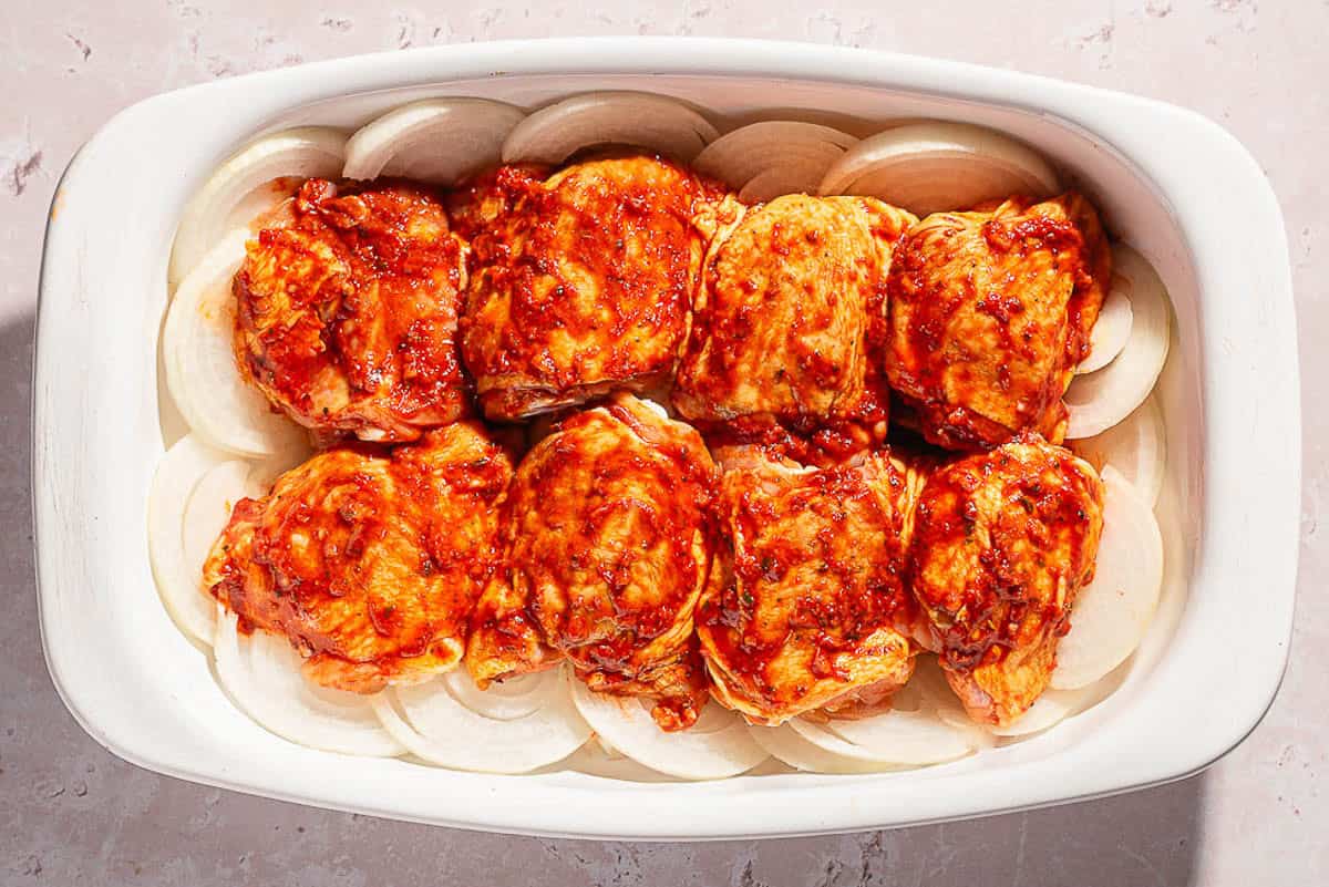 Unbaked chicken thighs topped with a tomato rub on a bed of sliced onions in a baking dish.