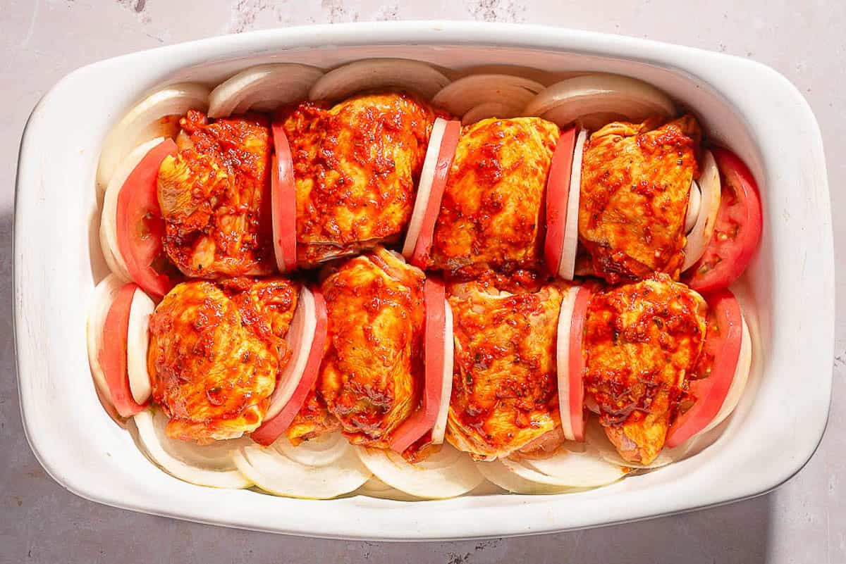 Unbaked chicken thighs separated with tomato and onion slices topped with a tomato rub on a bed of sliced onions in a baking dish.