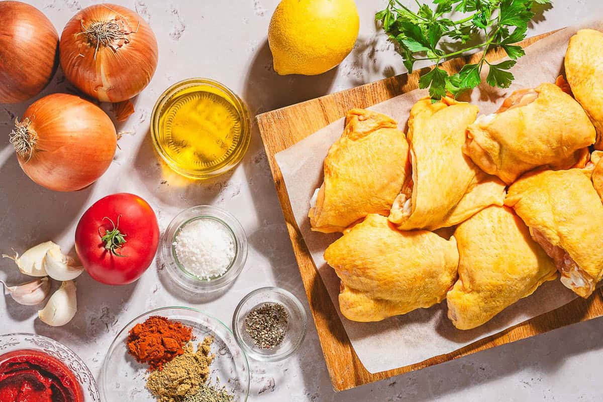 Ingredients for baked chicken thighs including bone in chicken thighs, salt, black pepper, olive oil, onions, a tomato, tomato paste, lemon, garlic, oregano, smoked paprika, and cumin.