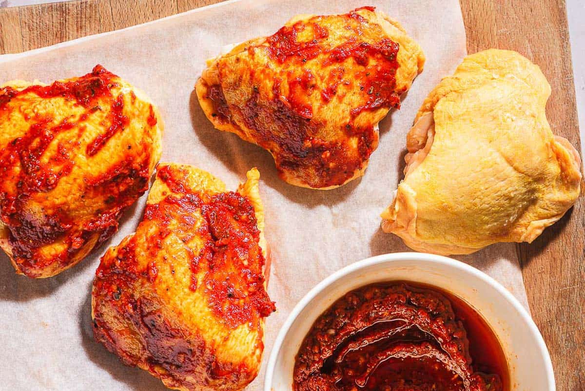 An overhead photo of 4 unbaked chicken thighs, 3 topped with a tomato rub, on a parchment lined cutting board. Next to these is a bowl of the rub.