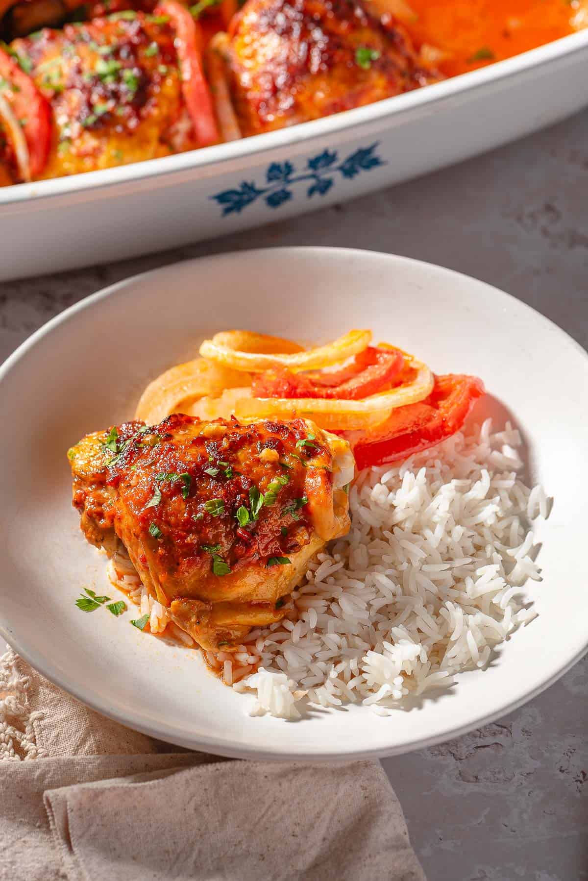 A close up of a baked chicken thigh with tomatoes and onion and a serving of rice on a plate. In the background is a baking dish with the rest of the chicken thighs.