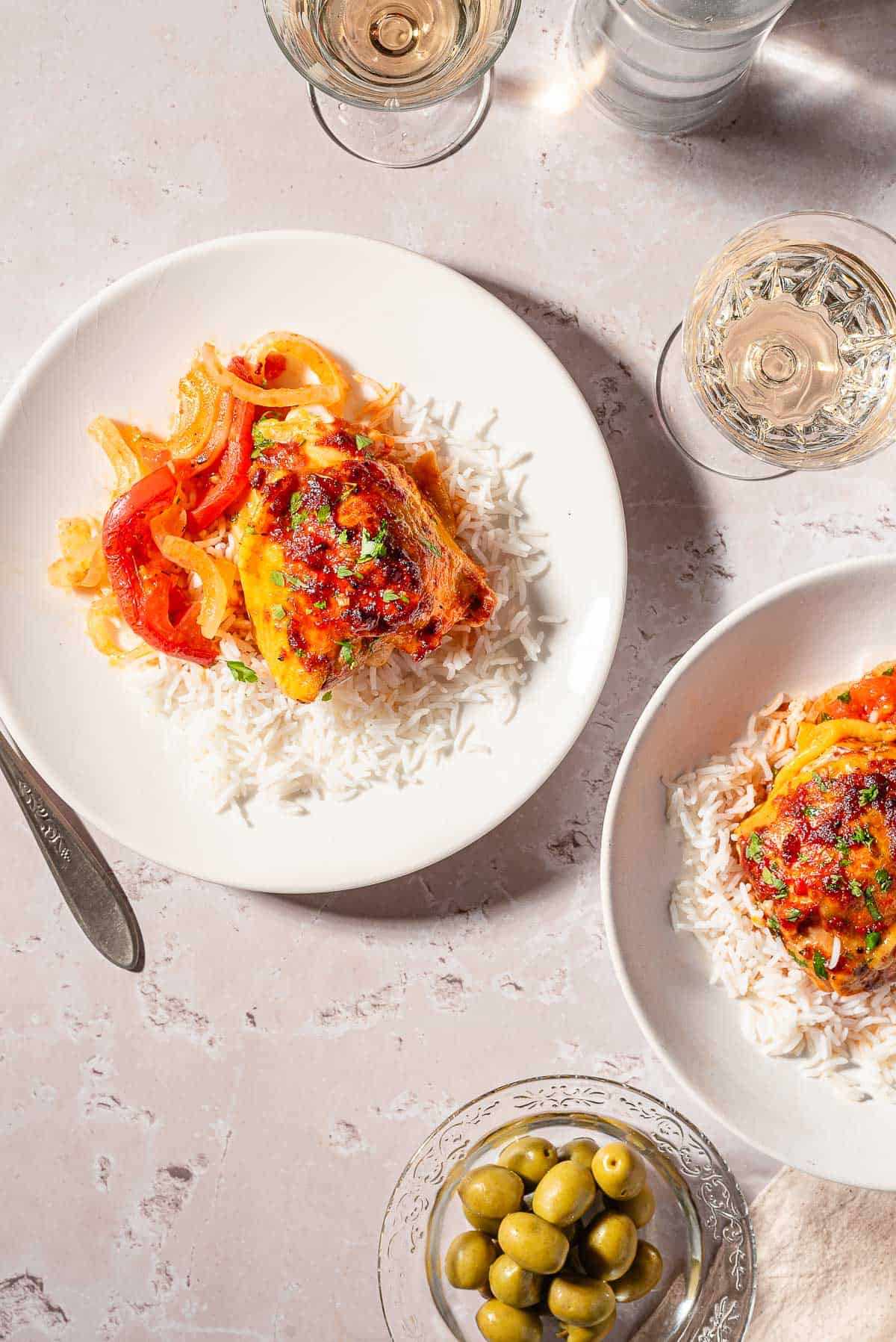 An overhead photo of a baked chicken thigh with tomatoes and onion and a serving of rice on a plate. Next to this is another plate with a chicken thigh and rice, 2 glasses of white wine, and a bowl of green olives.