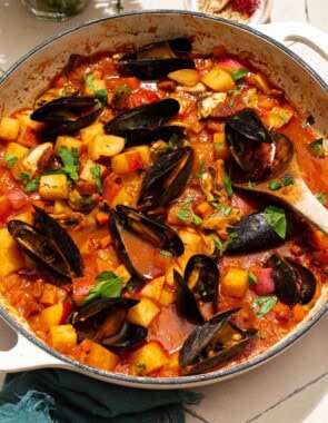 A close up of a pot mussels with chorizo in a saffron stew with a wooden spoon. Next to this a cloth napkin, bowls of spices, and some parsley.