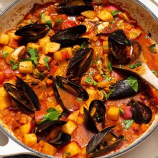 A close up of a pot mussels with chorizo in a saffron stew with a wooden spoon. Next to this a cloth napkin, bowls of spices, and some parsley.