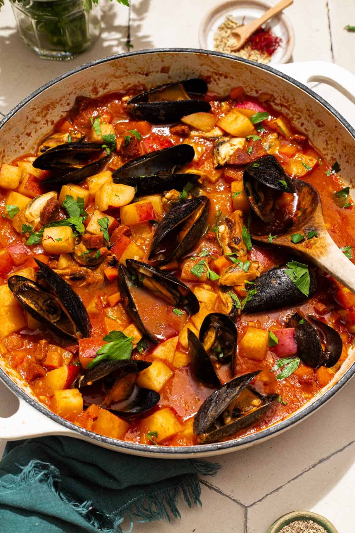 A close up of a pot mussels with chorizo in a saffron stew with a wooden spoon. Next to this a cloth napkin, bowls of spices, and some parsley.