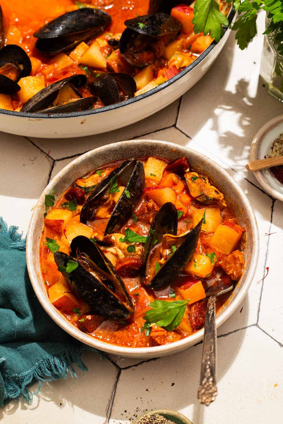 An overhead photo of a bowl of mussels with chorizo in a saffron stew with a fork. Next to this is the pot with the rest of the stew, a cloth napkin, bowls of spices, and some parsley.