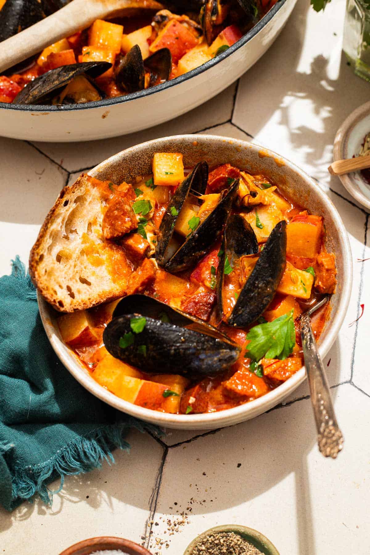 An overhead photo of a bowl of mussels with chorizo in a saffron stew with a fork and a slice of crusty bread. Next to this is the pot with the rest of the stew, a cloth napkin, and bowls of spices.