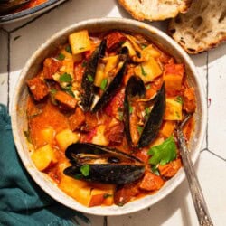 An overhead photo of a bowl of mussels with chorizo in a saffron stew with a fork. Next to this is the pot with the rest of the stew, a cloth napkin, bowls of spices, and slices of crusty bread.