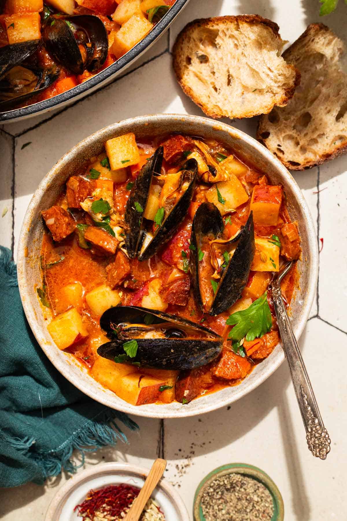 An overhead photo of a bowl of mussels with chorizo in a saffron stew with a fork. Next to this is the pot with the rest of the stew, a cloth napkin, bowls of spices, and slices of crusty bread.