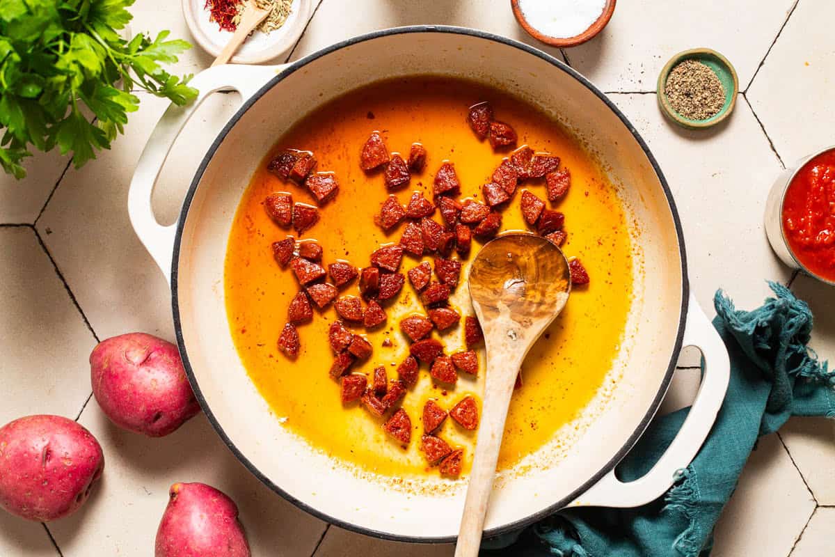 An overhead photo of chopped chorizo being sauteed in olive oil in a large pot with a wooden spoon. Next to this is a cloth napkin, parsley, a can of crushed tomatoes, red potatoes and bowls of spices.