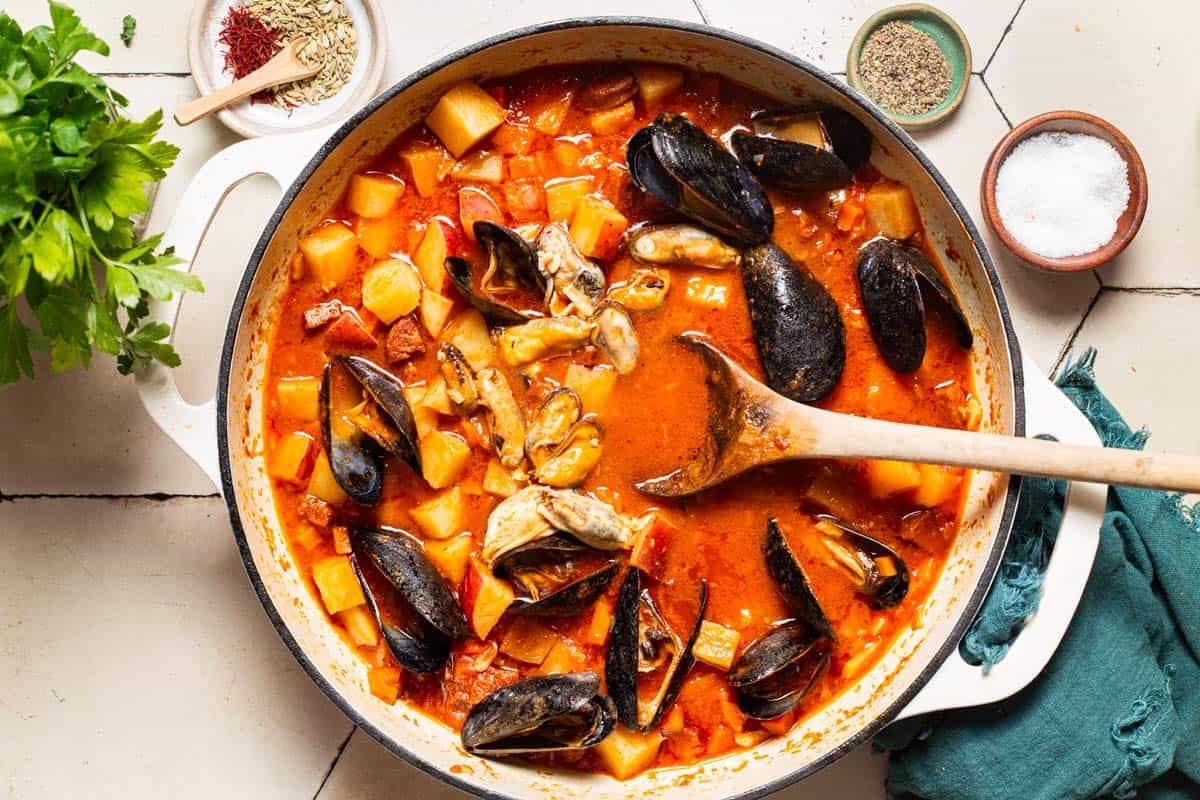 An overhead photo of a pot mussels with chorizo in a saffron stew with a wooden spoon. Next to this a cloth napkin, bowls of spices, and some parsley.