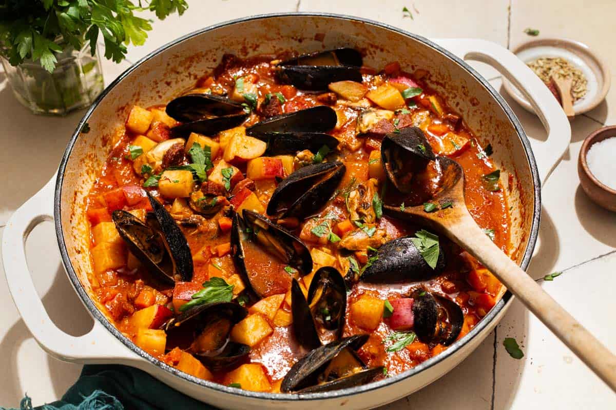 A close up of a pot mussels with chorizo in a saffron stew with a wooden spoon. Next to this a cloth napkin, bowls of spices, and some parsley.