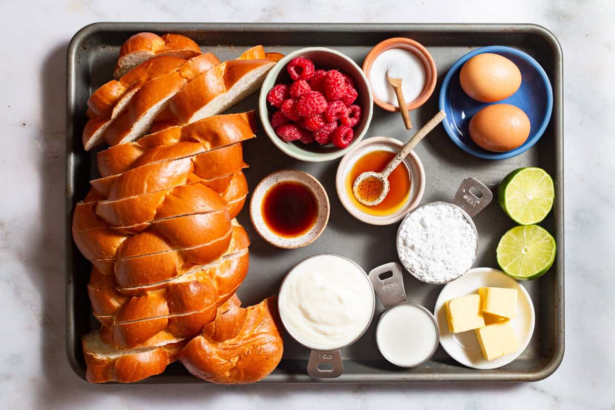 Ingredients for baked french toast including challah bread, plain yogurt, butter, eggs, honey, vanilla extract, salt, milk, powdered sugar, raspberries, and lime.