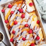 An overhead photo of baked french toast topped with powdered sugar and raspberries and drizzled with honey simple syrup in a baking dish.