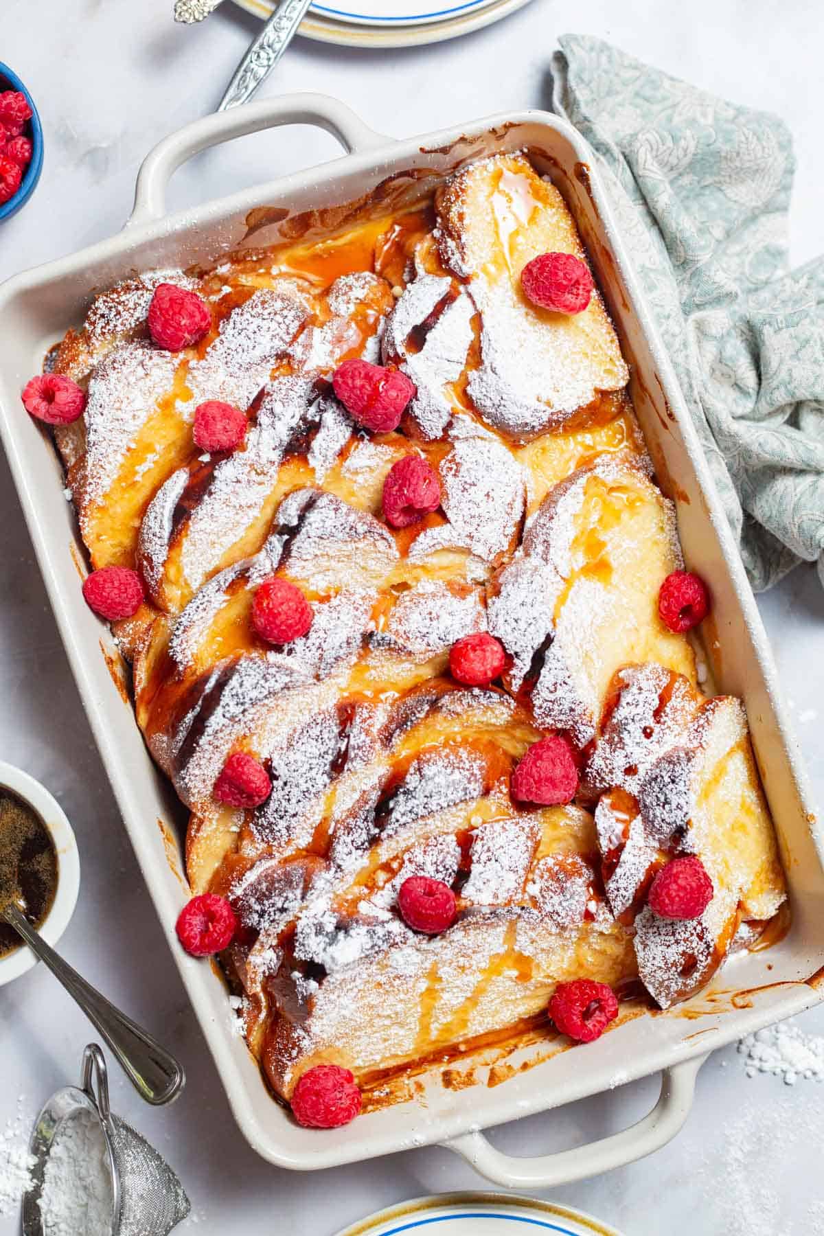 An overhead photo of baked french toast topped with powdered sugar and raspberries and drizzled with honey simple syrup in a baking dish.