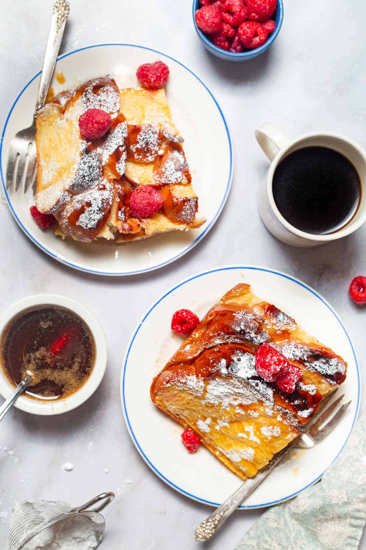 An overhead photo of 2 plates of baked french toast topped with raspberries and powdered sugar, and drizzled with honey. Next to these is a cup of coffee, a bowls of raspberries and a bowl of the simple syrup with a spoon.