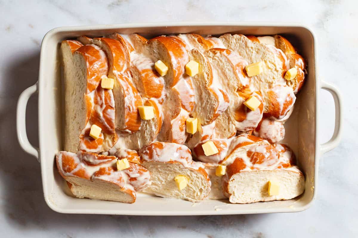 An overhead photo of slices of challah arranged in a baking dish topped with the custard and several pats of butter.