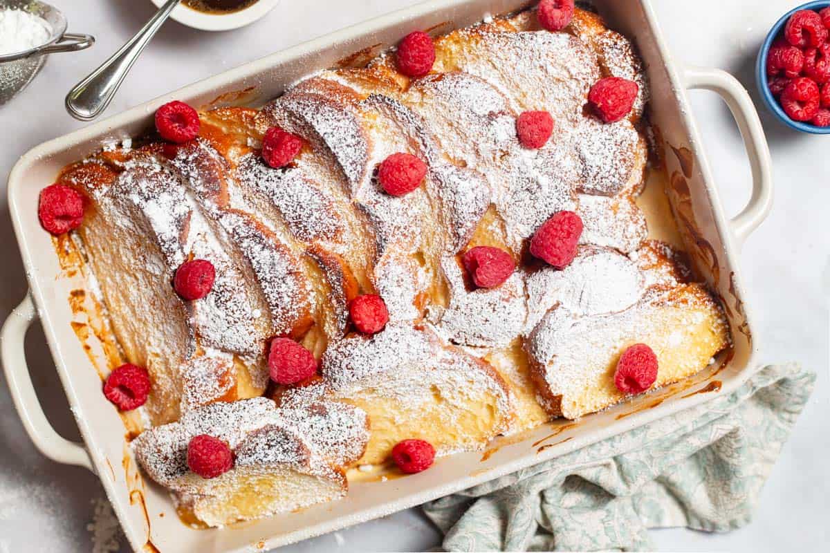 An overhead photo of baked french toast topped with powdered sugar and raspberries and drizzled with honey simple syrup in a baking dish. Next to this is a kitchen towel and a bowl of raspberries.