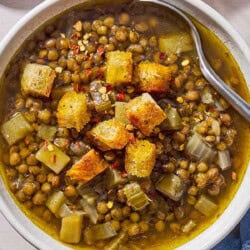 An overhead photo of a bowl of lentil potato soup with a spoon, topped with croutons. Next to this is a spoon, a cloth napkin, a bottle of olive oil, and a pot with the rest of the soup.