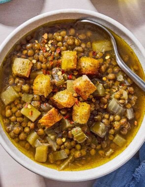 An overhead photo of a bowl of lentil potato soup with a spoon, topped with croutons. Next to this is a spoon, a cloth napkin, a bottle of olive oil, and a pot with the rest of the soup.