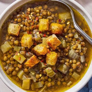 An overhead photo of a bowl of lentil potato soup with a spoon, topped with croutons. Next to this is a spoon, a cloth napkin, a bottle of olive oil, and a pot with the rest of the soup.